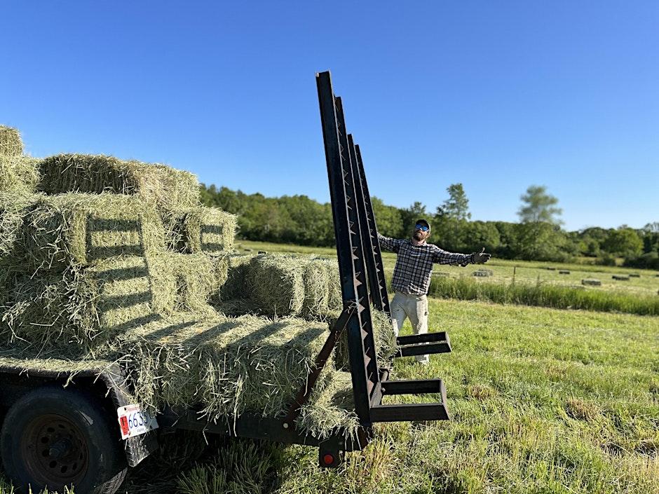 Owner of Freedom Food Farm standing next to a trailer full of hay with his arms outstretched
